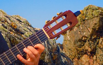 Close up fingers playing on the frets of a neck of a guitar, rocks and blue sky are in the background.
