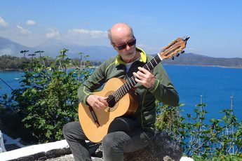 A man playing an acoustic guitar outside, the sea, foliage and hills are in the background.