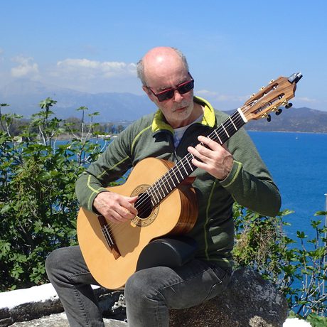 A man playing an acoustic guitar outside, the sea, foliage and hills are in the background.