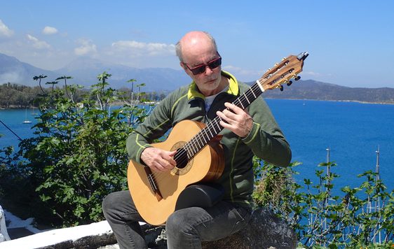 A man playing an acoustic guitar outside, the sea, foliage and hills are in the background.