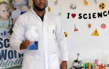 A person in a lab coat holding a beaker of blue smoking liquid, alongside a table of beakers with colourful liquid and sign that reads 'I love science'.
