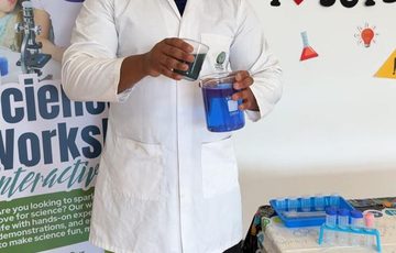 A person wearing a lab coat standing while mixing two glass beakers with colourful liquids.