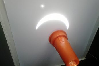 A crescent image of the Sun is projected onto a white screen through an orange tube holding a lens