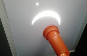 A crescent image of the Sun is projected onto a white screen through an orange tube holding a lens