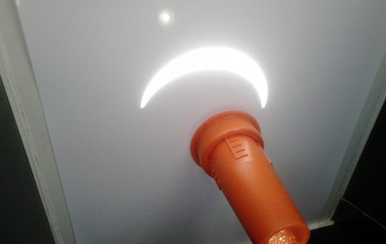 A crescent image of the Sun is projected onto a white screen through an orange tube holding a lens