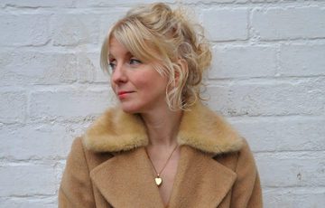 A lady looking away from the camera wearing a a coat and against a white brick wall.