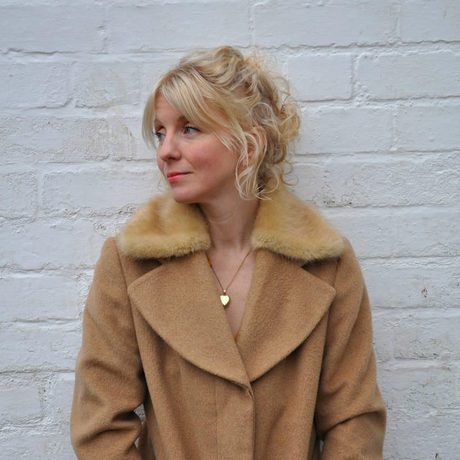 A lady looking away from the camera wearing a a coat and against a white brick wall.