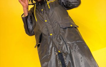 A person standing wearing a coat and hood, shot from below. A yellow background is behind.