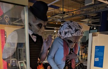 Two people dressed as alien Roswell characters looking at buttons on an interactive.