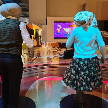 Back of two people dressed as alien Roswell characters, they are dancing in front of a Move It dance exhibit.