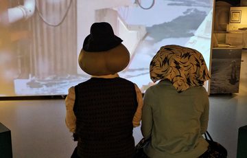 Back of heads of two people dressed as alien Roswell characters, they are watching a large screen.