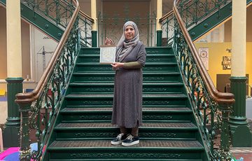 A person holding an artwork on an ornate metal staircase in a gallery.
