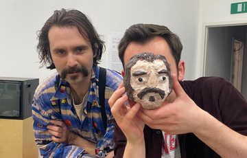 The artist with an adult who is holding their artwork (a face) to cover their own face.