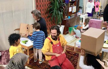 An artist workshop in studio with tables and art materials, the artist is talking to children and adults.