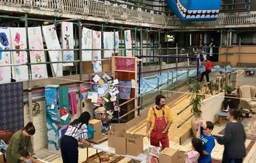 A wide shot of an artist workshop in studio, children and adults are taking part. Artworks surrounds the walls including a large blue square sea creature.