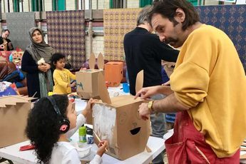 An artist helping a young child with a cardboard box craft, other people are in the background in the workshop.