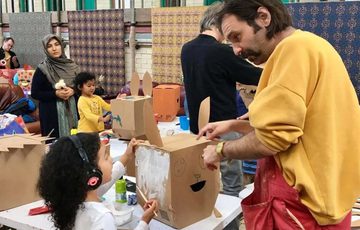 An artist helping a young child with a cardboard box craft, other people are in the background in the workshop.