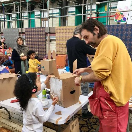 An artist helping a young child with a cardboard box craft, other people are in the background in the workshop.