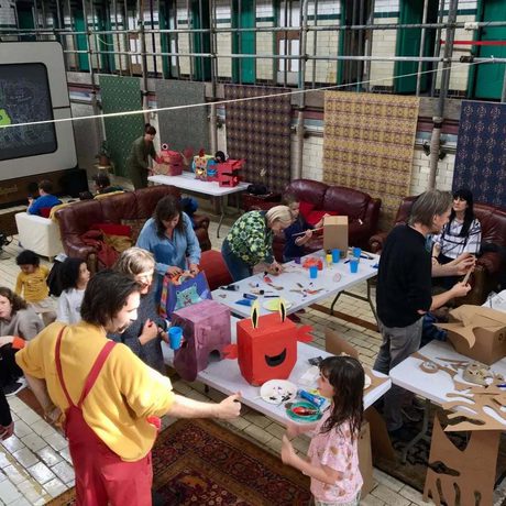 An artist workshop in studio with tables and art materials, children and adults are taking part.