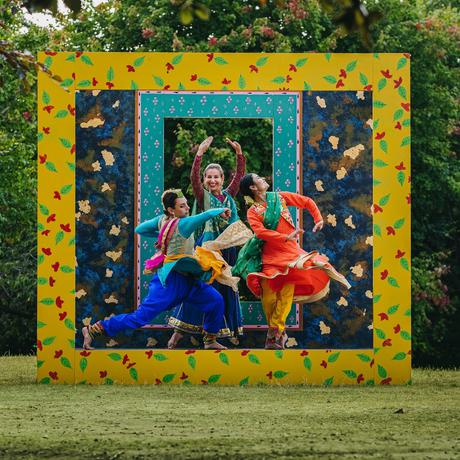 Three dancers performing outside with square and rectangular frames behind them as they jump in the air.