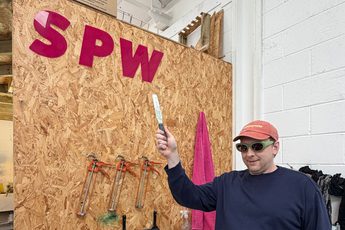 Joe Lycett pointing to a sign that reads 'S P W' in an indoor studio.