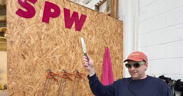 Joe Lycett pointing to a sign that reads 'S P W' in an indoor studio.