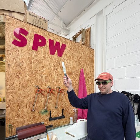 Joe Lycett pointing to a sign that reads 'S P W' in an indoor studio.
