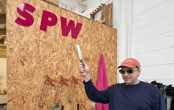 Joe Lycett pointing to a sign that reads 'S P W' in an indoor studio.