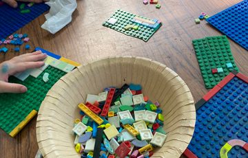 A table with hands holding lego boards and a bowl of LEGO pieces in the centre.