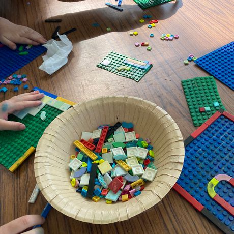 A table with hands holding lego boards and a bowl of LEGO pieces in the centre.