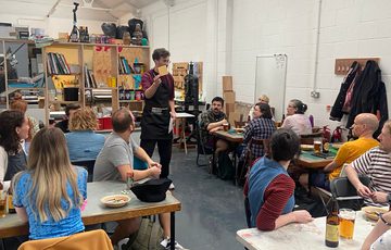 A workshop space with tables and people looking at a person who is standing up and presenting.