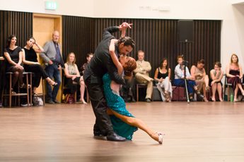 Two professional dancers performing a dance together, people are sat around watching.