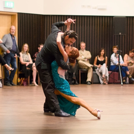 Two professional dancers performing a dance together, people are sat around watching.