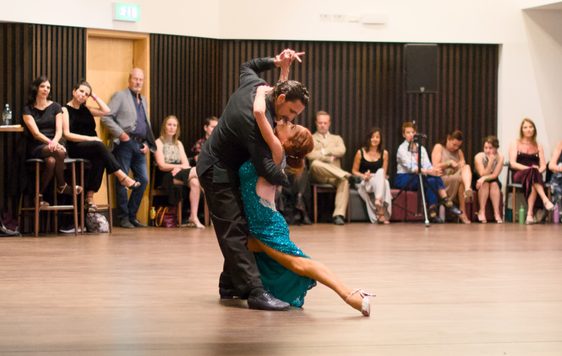 Two professional dancers performing a dance together, people are sat around watching.