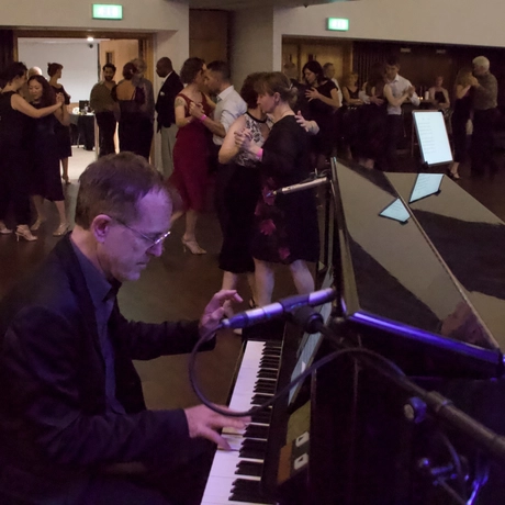 A pianist playing a piano while a group of people dance in pairs.