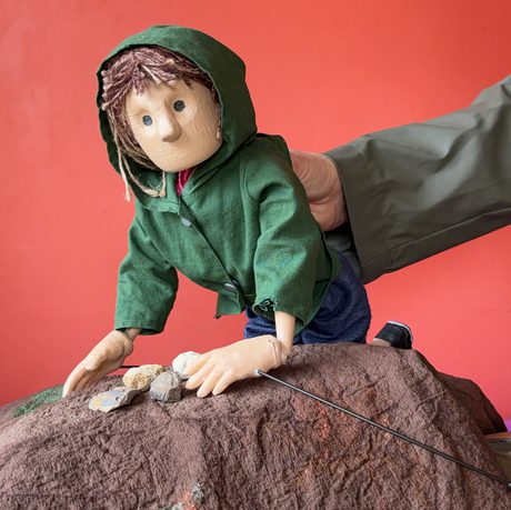 A close up of a hand holding a puppet of a person leaning on a rock.