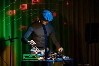 A person performing with equipment on a desk and coloured lights behind.