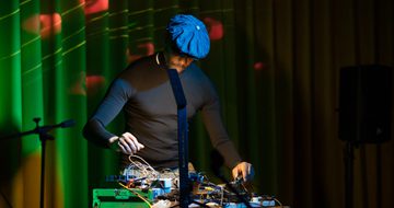 A person performing with equipment on a desk and coloured lights behind.