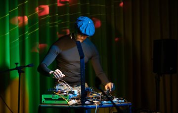 A person performing with equipment on a desk and coloured lights behind.