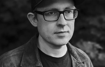 A black and white portrait of a person wearing glasses and a cap.