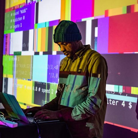 A person with computer in front of them and pixel coloured screen behind.