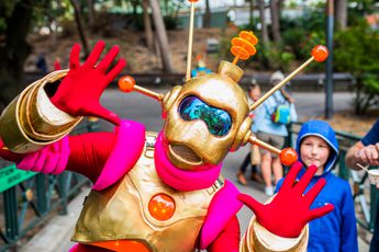 An alien characters with red, gold and pink outfit posing with their hands up, a child is looking behind.