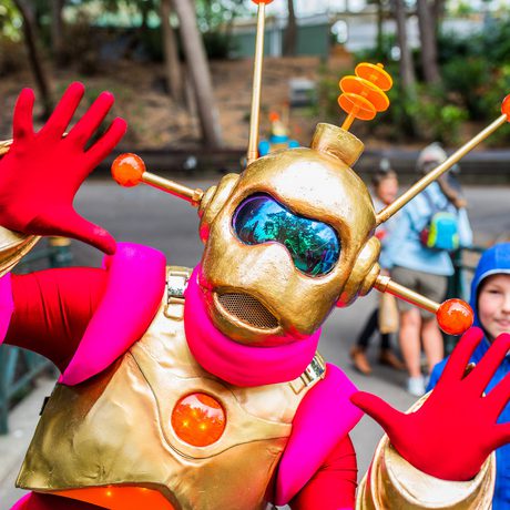 An alien characters with red, gold and pink outfit posing with their hands up, a child is looking behind.