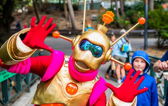 An alien characters with red, gold and pink outfit posing with their hands up, a child is looking behind.