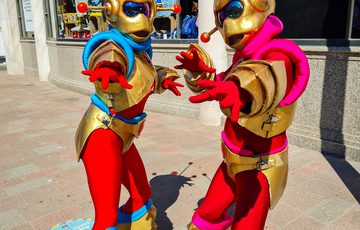 Two alien characters with red and gold outfits outside on street, posting for the camera with their arms out.