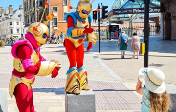 Two alien characters with red and gold outfits, on a street with a small child looking at them.