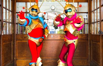 Two alien characters with red and gold outfits, inside a wooden room with windows and chandelier.