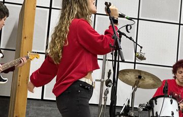 A lady wearing a red top and black jeans singing on a microphone with drummer and guitarist partially shown in the background.