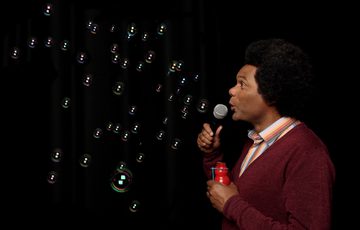Andy White performing holding a microphone and looking at bubbles against a black background.
