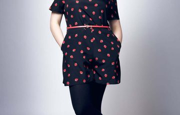 Lasy with red hair wearing a dark blue dress and red shoes with hands in pockets, standing against a light grey background